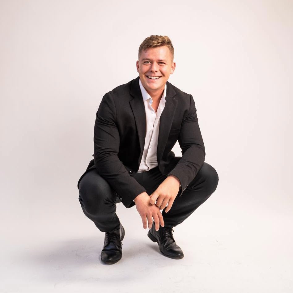 Studio portrait of Matt Fitzsimmons in a dark suit and white shirt, crouching and smiling.